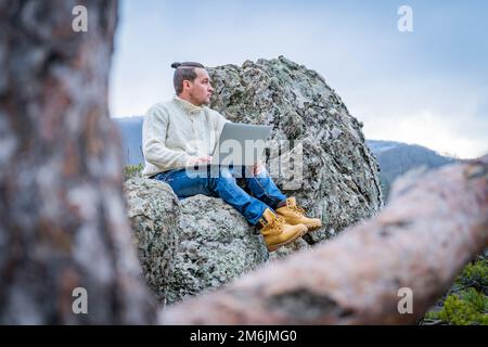 Hipster man working with laptop sitting on the rocky mountain on beautiful scenic clif background Stock Photo