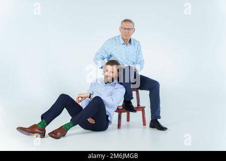 Father sits on the chair and son sits on white background, both men look into the camera, an elderly man in glasses with diopter Stock Photo
