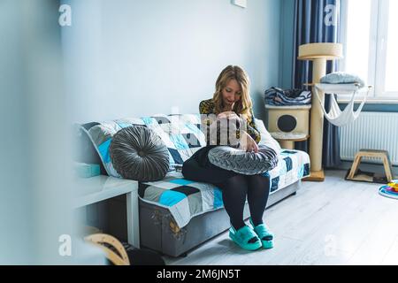 A woman sitting on a couch in a room and holding a Devon rex cat in her hands. Cat equipment in the background. Wide shot. High quality photo Stock Photo