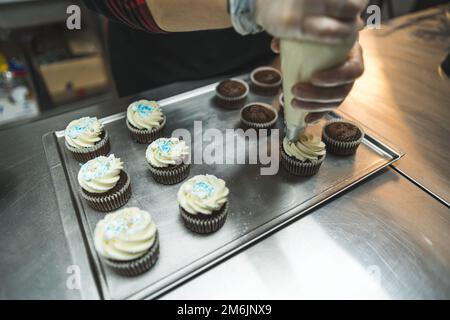 Cupcakes placed on a baking tray being decorated with white cream. Six cupcakes decorated with cream and blue sprinkles. A baker wearing transparent gloves decorating cupcakes with a pastry bag. Blurred foreground. High quality photo Stock Photo