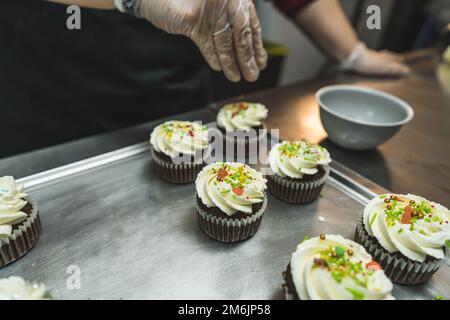 A baker wearing a transparent gloves decorating cupcakes on a baking tray. Cupcakes with white cream being sprinkled with colorful flakes. Blurred background. . High quality photo Stock Photo