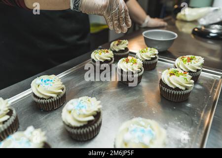 Side view of six cupcakes decorated with white cream being sprinkled with colorful flakes. Blurred cupcakes decorated with white cream and blue sprinkles in the foreground. A person wearing a transparent glove decorating cupcakes. High quality photo Stock Photo