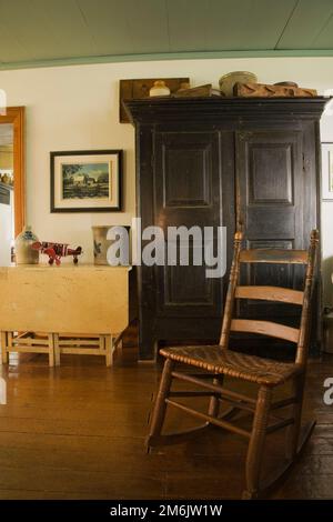 Old wooden antique weaved seat rocking chair and armoire in dining room inside old 1785 fieldstone cottage style home. Stock Photo