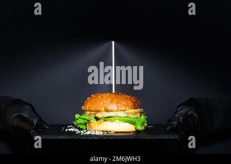 Savoury cheeseburger on a plate holding in hands in black glooves. On a black background. Stock Photo