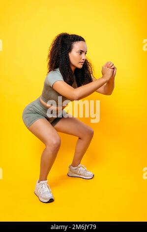 Sports lifestyle. Full length vertical photo of a fitness sporty lovely curly haired brazilian or latino woman in sportswear, practicing squat exercise on isolated orange background Stock Photo