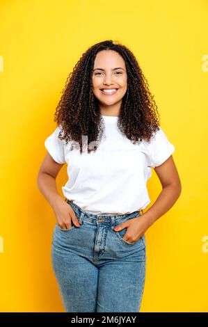 Vertical photo of lovely positive cheerful brazilian or hispanic young curly haired woman, in casual basic clothes, happily smiling at camera, stands over isolated orange background, hands in pockets Stock Photo