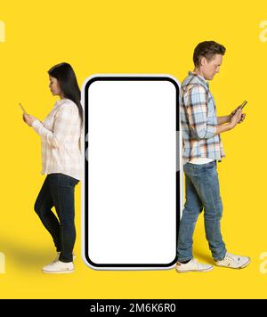 Asian woman and caucasian man standing leaned on huge smartphone with white screen looking at phones in hands, mobile app advert Stock Photo