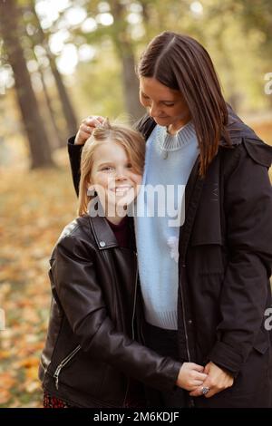 Portrait of glad family of young woman and teenage girl standing on yellow fallen leaves in forest park in autumn. Stock Photo