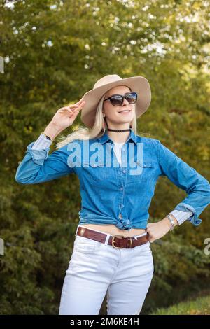 Portrait of young wonderful blond woman touching beige floppy hat, standing near green trees forest park. Travelling. Stock Photo
