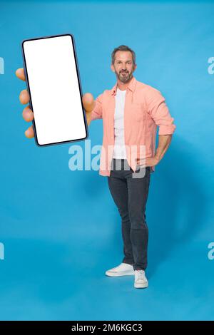 Grey haired middle aged man gesturing hello with one hand up happy smiling on camera wearing peach color shirt and grey jeans is Stock Photo