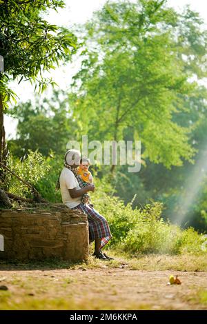 NOVEMBER 15 2022, KACHANDA, CHHATTISGARH, INDIA: Grandfather holding grandson in indian village and sitting under the tree in morning sunlight. Grandf Stock Photo