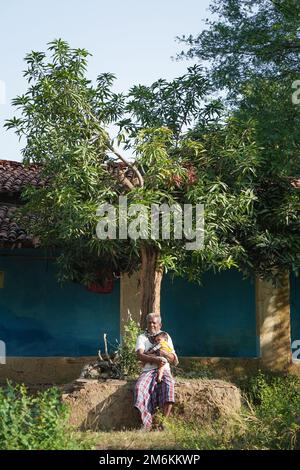 NOVEMBER 15 2022, KACHANDA, CHHATTISGARH, INDIA: Grandfather holding grandson in indian village and sitting under the tree in morning sunlight. Grandf Stock Photo