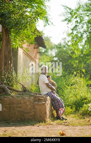 NOVEMBER 15 2022, KACHANDA, CHHATTISGARH, INDIA: Grandfather holding grandson in indian village and sitting under the tree in morning sunlight. Grandf Stock Photo