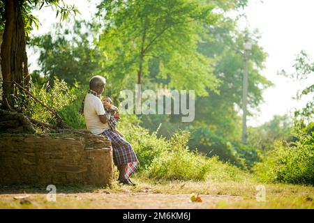 NOVEMBER 15 2022, KACHANDA, CHHATTISGARH, INDIA: Grandfather holding grandson in indian village and sitting under the tree in morning sunlight. Grandf Stock Photo