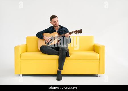 Handsome young man playing acoustic guitar whlile sitting on yellow sofa over white background Stock Photo