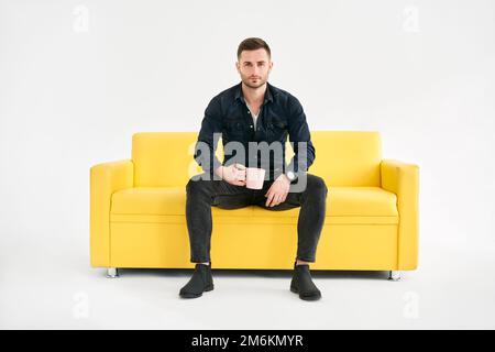 Relaxed man with coffee mug in hands sitting on comfort bright sofa looking to camera Stock Photo
