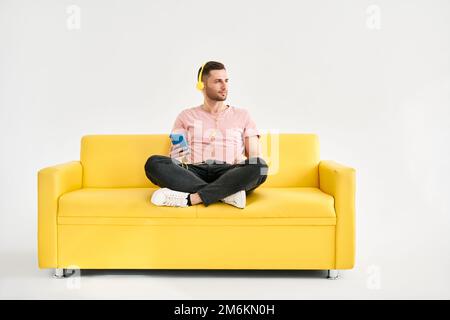 Relaxed young man wear headphones listen music by his mobile phone while sitting on yellow sofa Stock Photo