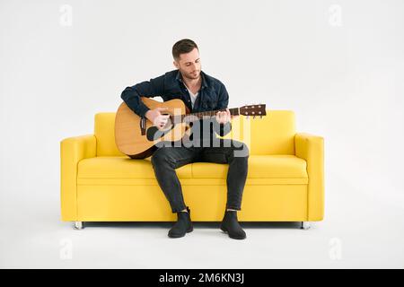 Handsome young man playing acoustic guitar whlile sitting on yellow sofa over white background Stock Photo
