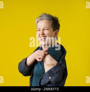 Grey hair senior woman showing shoulder with band aid after having shot of vaccine wearing trendy clothes isolated on yellow bac Stock Photo