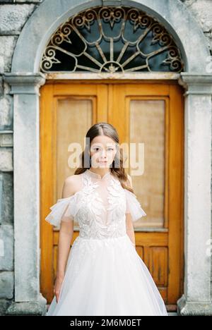 Bride in a white dress stands in front of a wooden door of a stone building Stock Photo