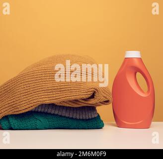 Stack of washed folded clothes and plastic orange large bottle with liquid detergent stand on a white table, yellow background. Stock Photo