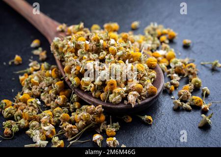 Traditional dried chamomile blossoms offered as close-up on a rustic wooden spoon on a black board Stock Photo
