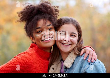 Two happy multiracial girlfriends smiling at camera and hugging while standing in autumn forest Stock Photo