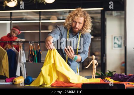 Young caucasian male fashion designer photographing fabric with smart phone in creative office Stock Photo