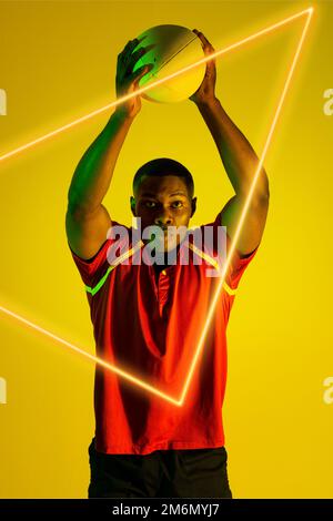African american male rugby player throwing ball over illuminated triangle on yellow background Stock Photo