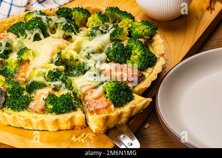 Closeup view of freshly baked delicious homemade broccoli and salmon quiche and the slice with white ceramic milk pot on wooden cutting board. Stock Photo