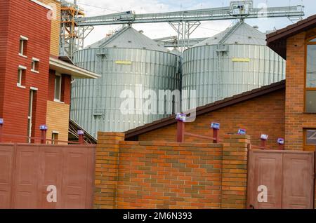 Granary among the houses. Neighborhood of a grain storage with a townhouse. Industry and household. Agricultural Silos. Storage and drying of grains, Stock Photo