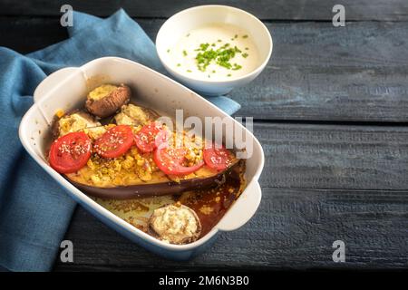 Baked half eggplant and stuffed mushrooms with garlic, ginger, onions and tomatoes in a casserole, served with yoghurt dip on a blue napkin and a dark Stock Photo