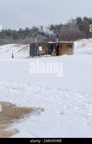 Wooden outdoor sauna with a smoke coming out of chimney on a beautiful cold snowy winter day at the Baltic sea. Well being and healthy life style. Stock Photo