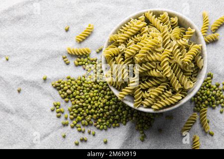 Mung bean fusilli pasta on a gray textile background. Bowl with raw pasta and green mung bean. Gluten free pasta. Stock Photo