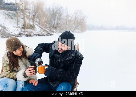 Happy lovely middle-aged couple in warm clothes drinking hot tea during romantic winter date in nature Stock Photo