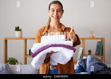 Thumbs up, laundry and portrait of a woman maid folding clothes in the living room in a modern house. Happy, smile and female cleaner or housewife Stock Photo