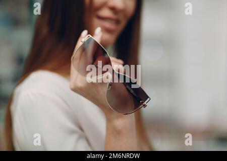 Close up of gorgeous young smiling woman smiling picking and choosing glasses at the optician corner at the shopping mall. Happy Stock Photo