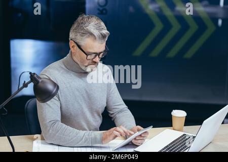 Serious thinking gray-haired businessman working inside office with laptop, senior man using tablet computer browsing internet pages reading online news. Stock Photo