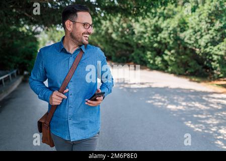 Delighted adult man in blue shirt, holding his phone, walking through the park outside. Stock Photo