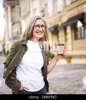 Happy mature businesswoman with disposable cup of hot coffee in hand. Charming senior woman traveling in Europe enjoying cup of Stock Photo