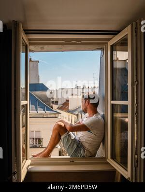 Young man sitting in a window looking out over the city Paris. Men enjoy sun in window of hotel room Stock Photo