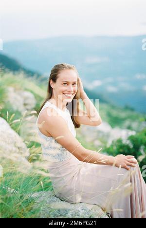 Smiling girl straightens her hair with her hand while sitting on a stone in the mountains Stock Photo