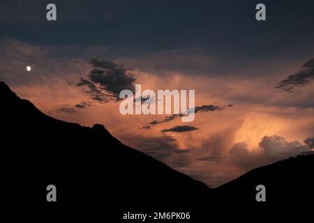 Beautiful sunset in valley between mountains. Sunset sky with orange clouds over hilly landscape. The moon at sunset sky above the mountains. Stock Photo