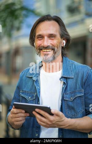 Confident freelancer man with grey haired hold digital tablet in hands having a call while standing on urban streets. Mature man Stock Photo