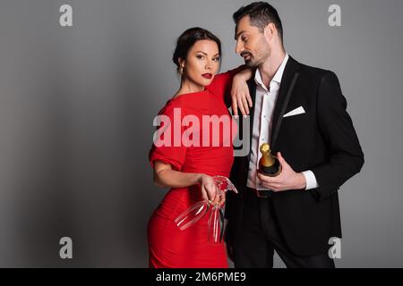 Man in formal wear holding champagne and looking at elegant girlfriend with glasses on grey background,stock image Stock Photo