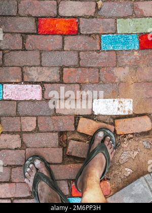 Feet of man wear sandals on old beautiful multicolor bricks paving stones footpath pattern on sidewalk outdoors as brickwork weave texture or abstract Stock Photo