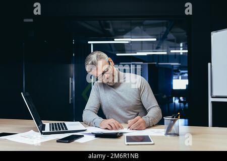 Senior mature financier investor working smiling and satisfied with documents, mature businessman on paper work with invoices and contracts using laptop inside office. Stock Photo