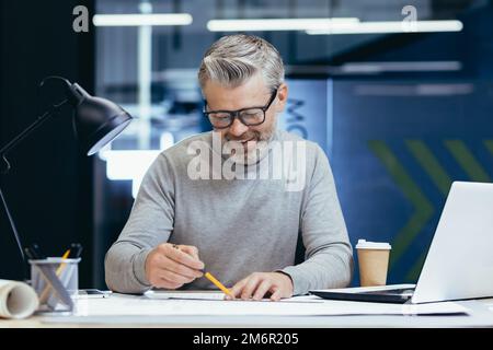 Successful senior architect at work in office, mature gray haired man drawing blueprints on layout and plan smiling and happy with work. Stock Photo