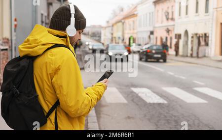 Man in yellow jacket and headphones standing next to road in city using his smart phone while waiting taxi. Stock Photo