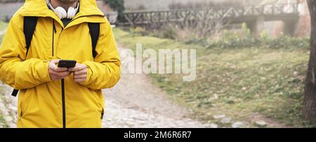 Man in yellow jacket with headphones on his neck and smart phone in hands walking outdoors. Stock Photo